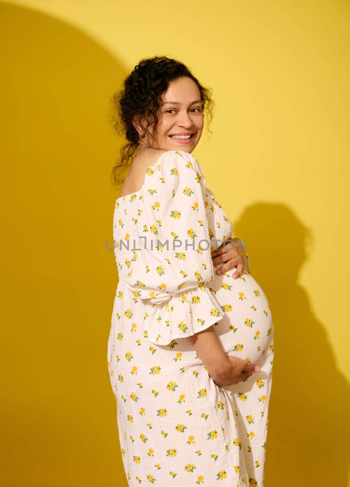 Curly multiethnic pregnant woman expecting a baby, smiling broadly looking at camera, isolated over yellow studio background. Childbearing. Childbirth. Beautiful pregnancy and maternity leave concept