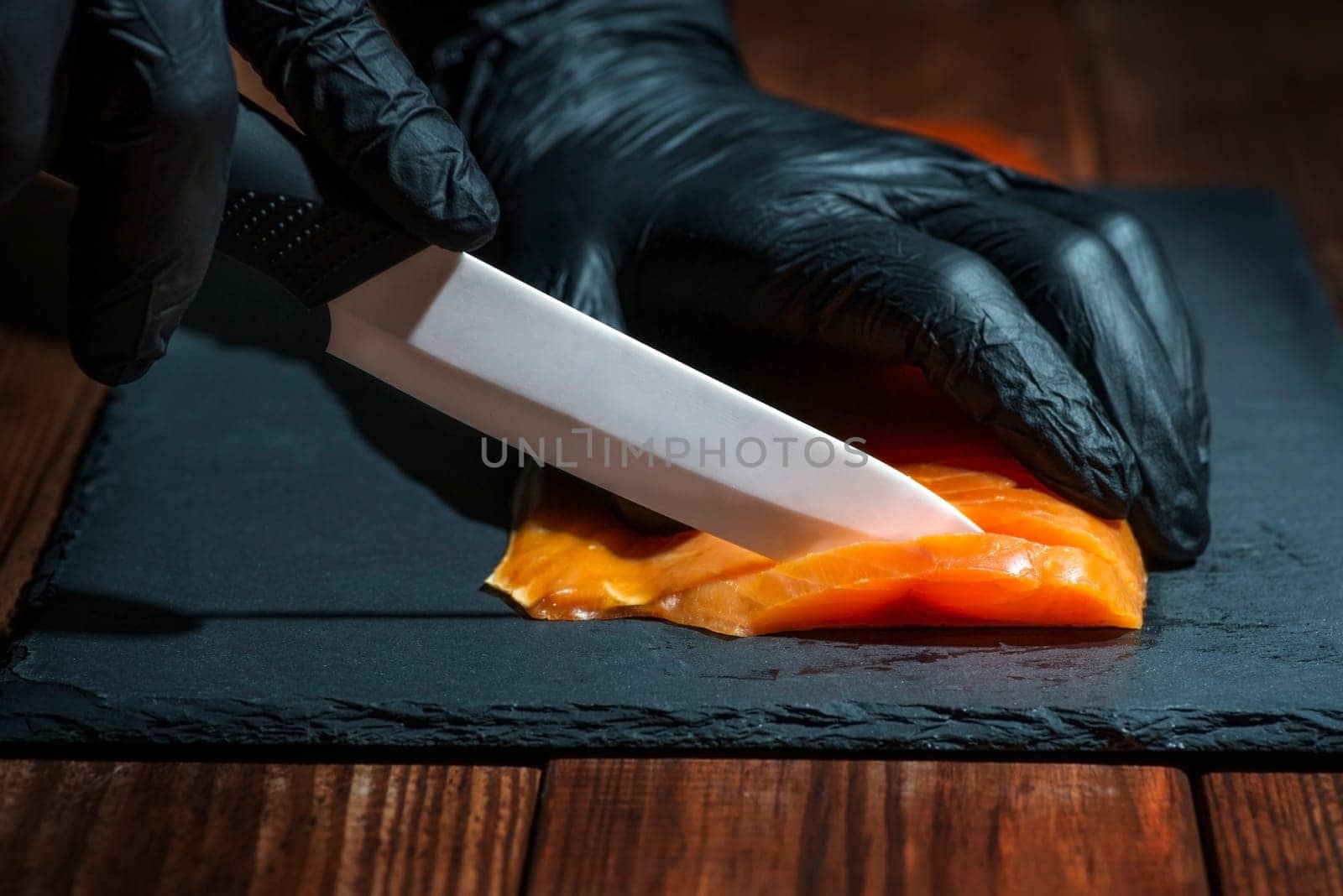 Chef use knife to cutting salmon fillet on black board at professional kitchen. Closeup chef hands in black gloves slicing red fish slice.