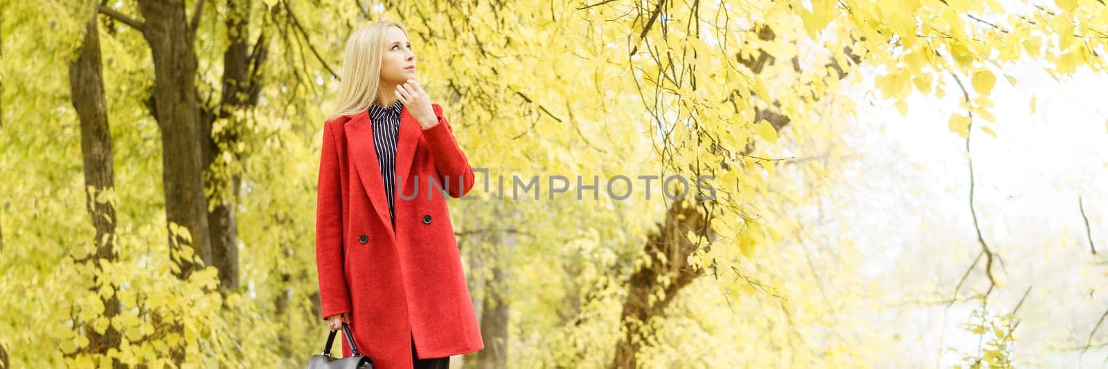 A young blonde woman walks around the autumn city in a red coat. The concept of urban style and lifestyle.