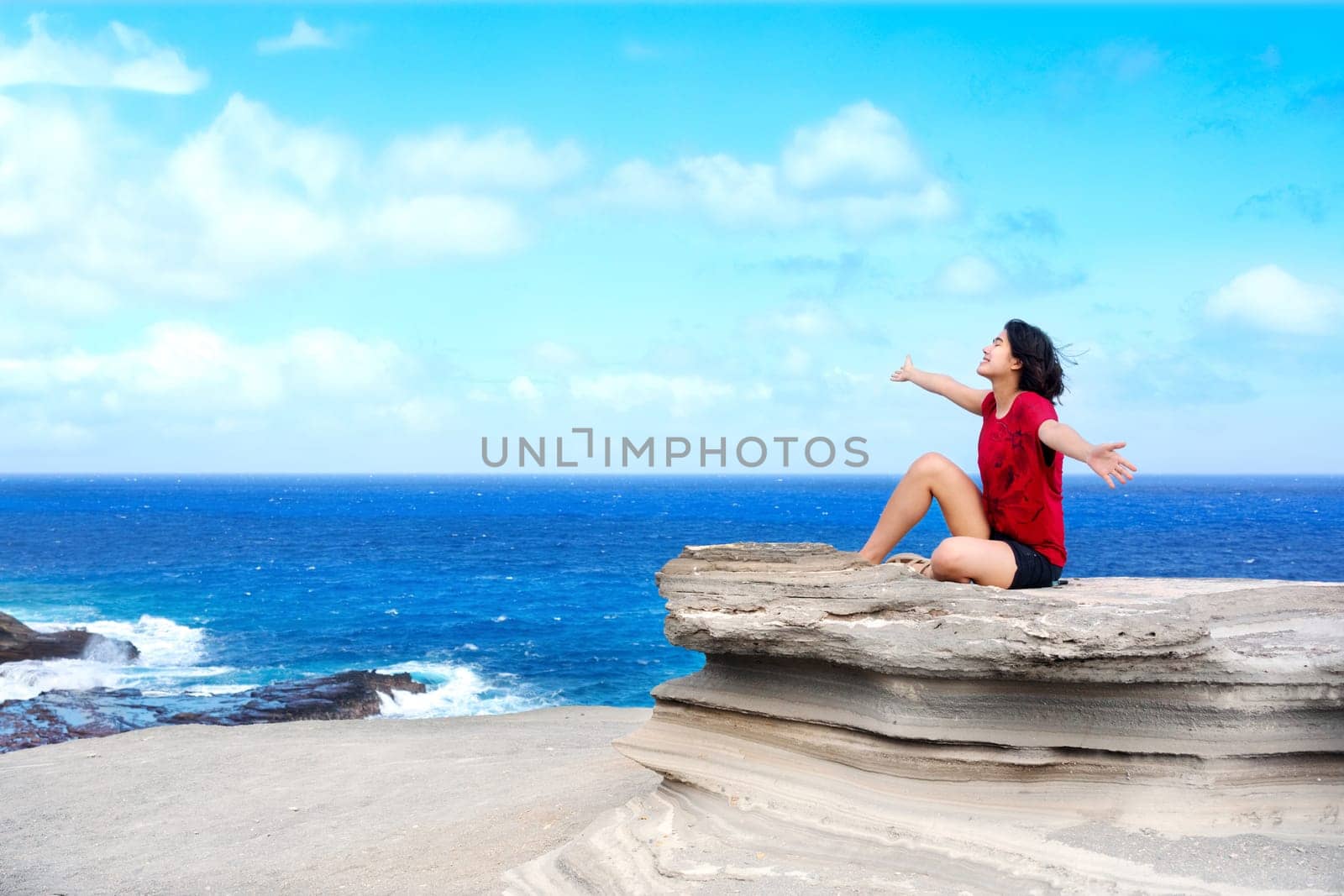 Young woman sitting on large rock cliff  by Hawaiian  ocean, arms raised enjoying breeze