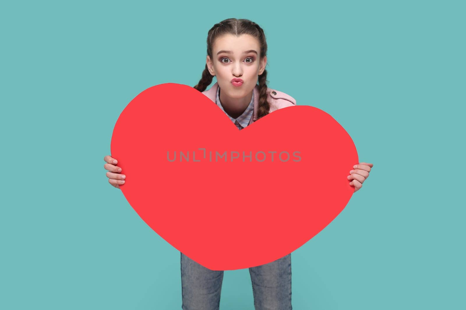 Portrait of optimistic romantic lovely teenager girl with braids wearing pink jacket holding big red heart, sending air kissing. Indoor studio shot isolated on green background.