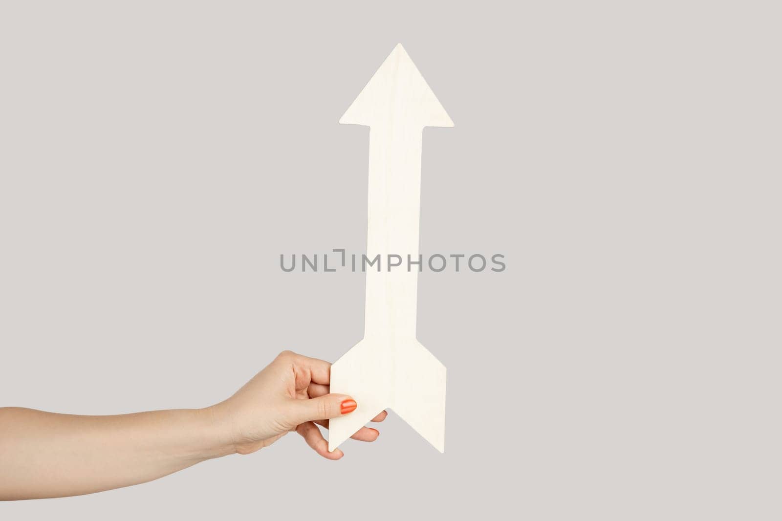 Closeup of woman hand holding white arrow indicating direction upwards, symbol of increase. Indoor studio shot isolated on gray background.