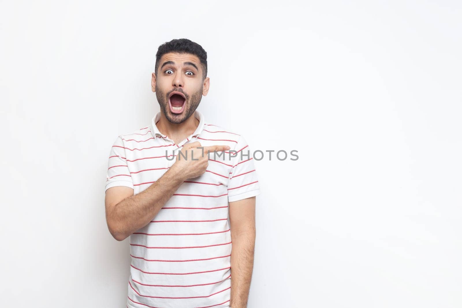 Portrait of astonished shocked bearded man wearing striped t-shirt standing pointing aside at copy space for promotion, advertisement area. Indoor studio shot isolated on gray background.