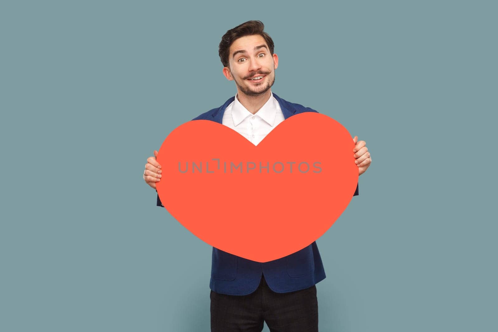 Portrait of positive excited amazed man with mustache standing holding big red heart, expressing romantic feelings, wearing white shirt and jacket. Indoor studio shot isolated on light blue background