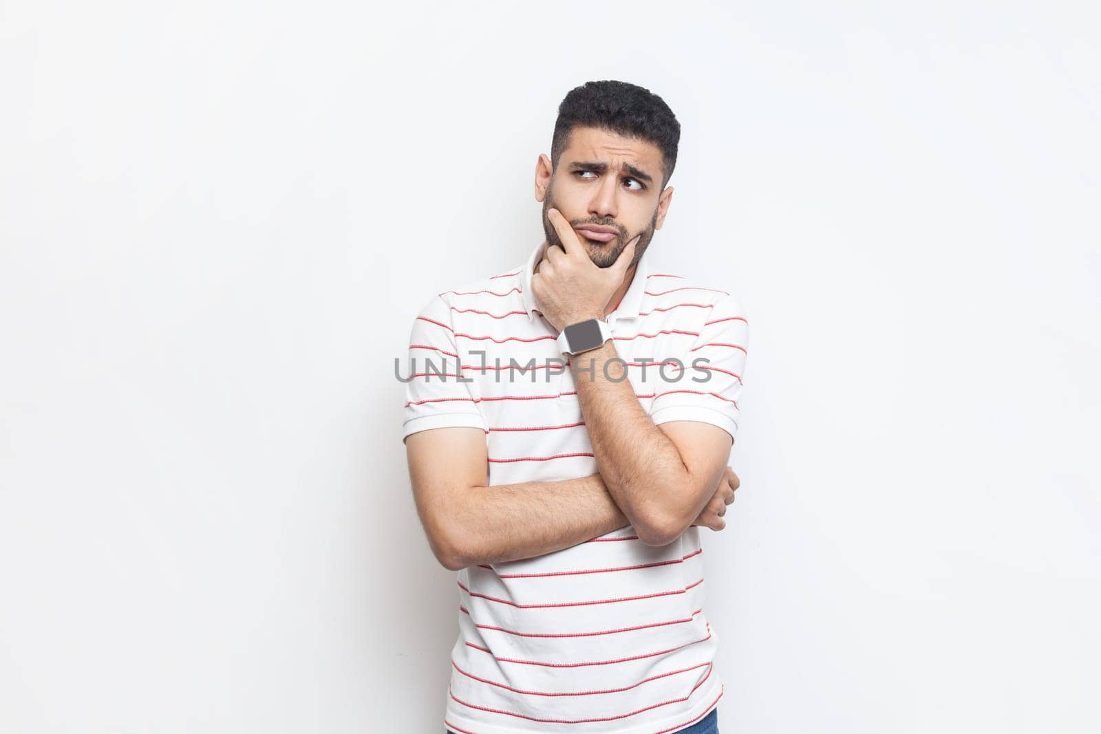 Portrait of pensive thoughtful attractive young adult bearded man wearing striped t-shirt standing holding chin, pondering, thinking. Indoor studio shot isolated on gray background.