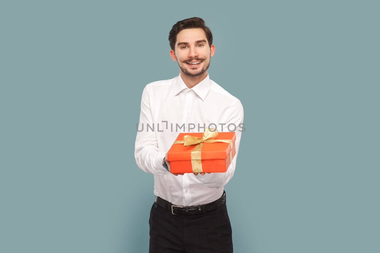 Portrait of friendly positive optimistic man with mustache giving red present box, congratulating with holiday, wearing Indoor studio shot isolated on light blue background.