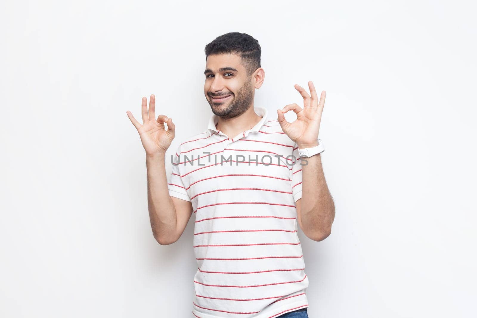 Everything ok. Portrait of attractive positive smiling bearded man wearing striped t-shirt standing showing okay gesture, agreed. Indoor studio shot isolated on gray background.