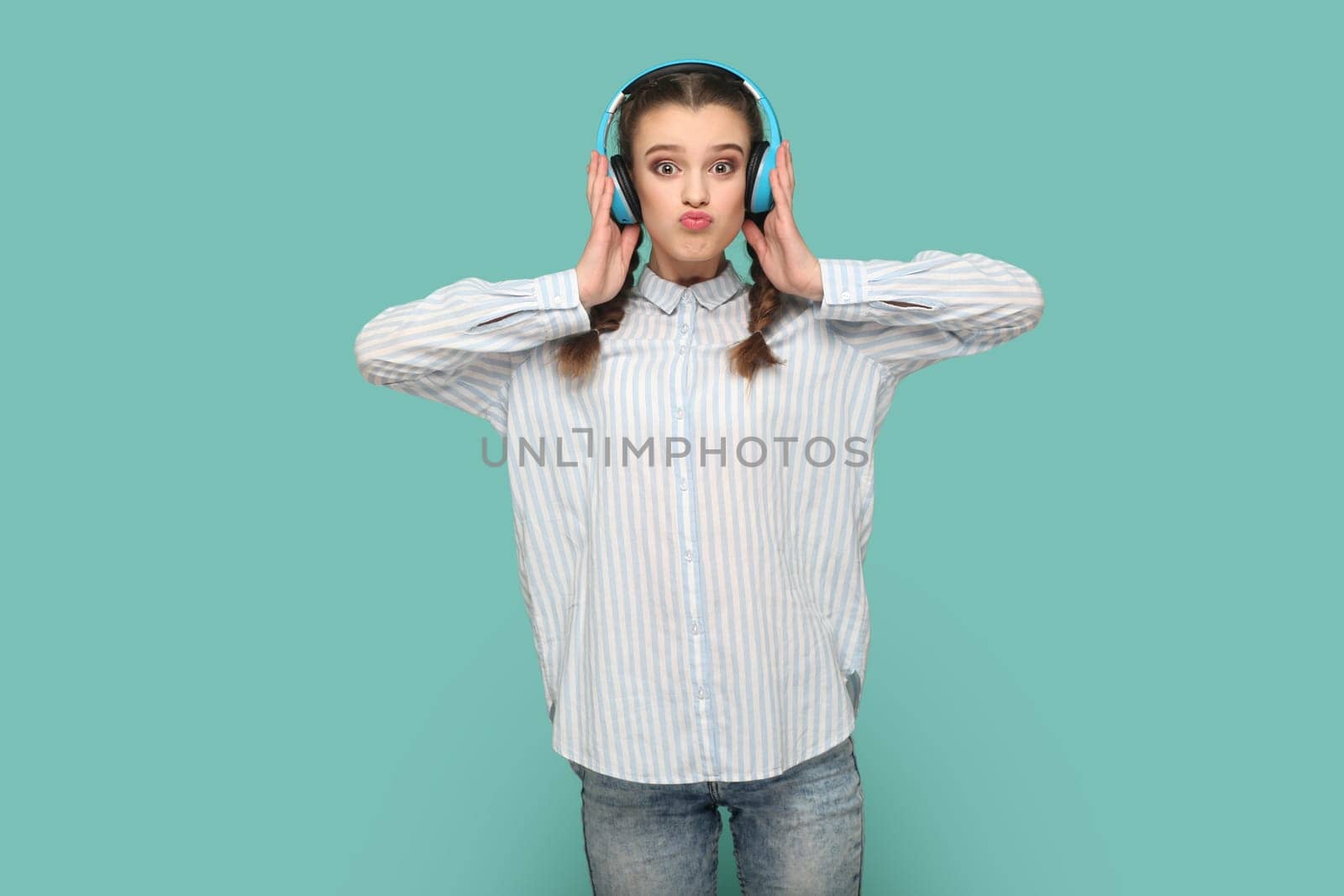 Portrait of funny teenager girl with braids wearing shirt standing looking at camera with pout lips, listening music, holding hands on headphones. Indoor studio shot isolated on green background.