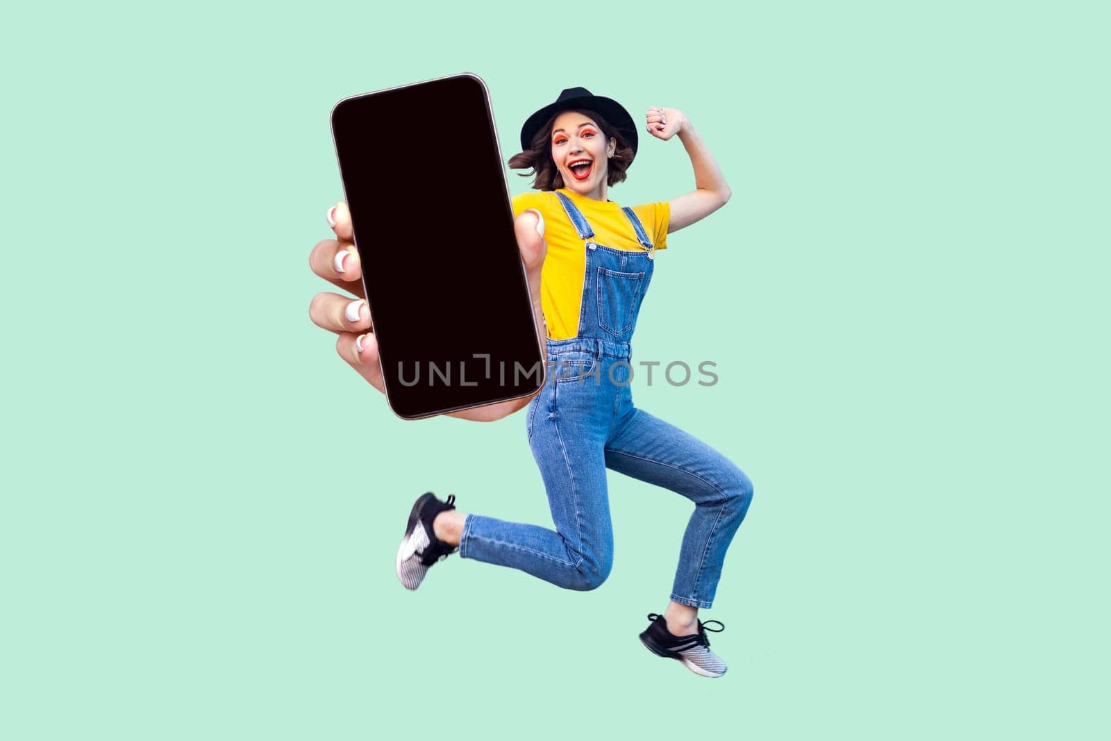 Portrait of happy surprised hipster girl in overalls, T-shirt and hat jumping and looking at camera, showing big empty smart phone display. Indoor studio shot isolated on light green background.