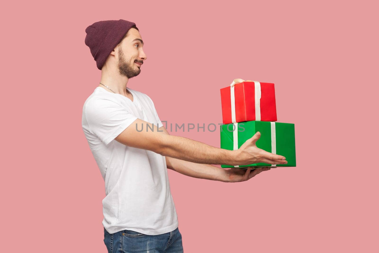 Side view portrait of smiling happy bearded man in white T-shirt and beany hat standing giving gift to his friend congratulating with birthday. Indoor studio shot isolated on pink background.