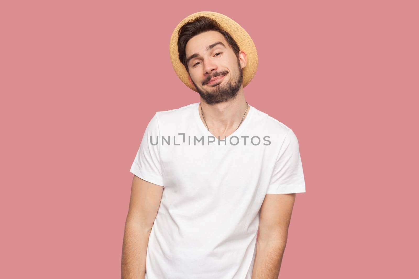 Portrait of cool self-confident smiling bearded man in white T-shirt and hat standing looking at camera, having satisfied expression. Indoor studio shot isolated on pink background.