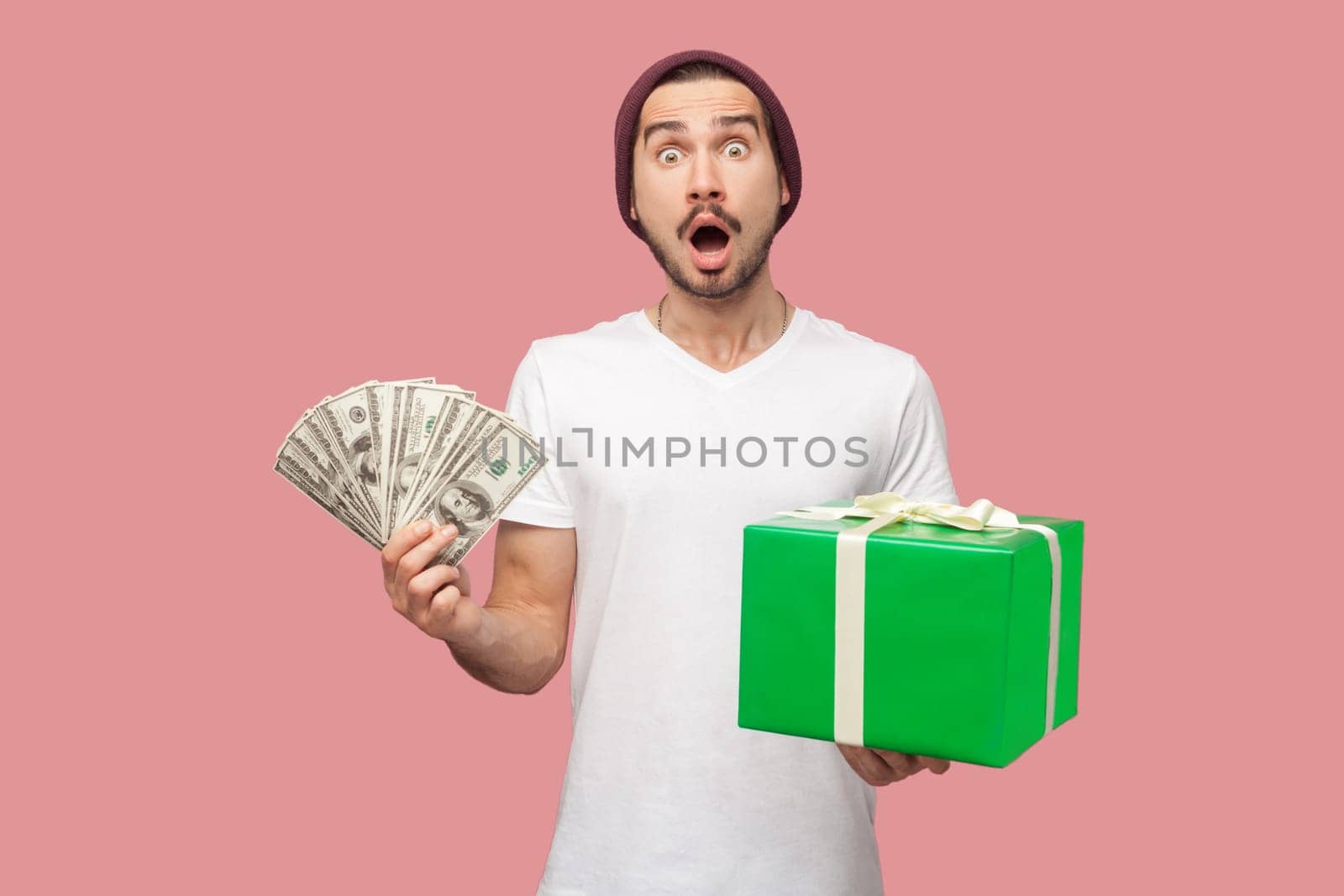 Portrait of shocked surprised man in white T-shirt and beany hat standing with dollar banknotes and green present box, looking at camera with open mouth. Indoor studio shot isolated on pink background