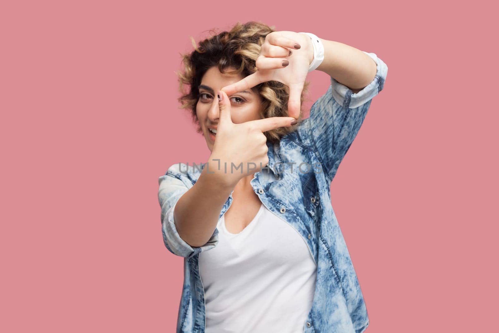 Portrait of concentrated attractive woman photographer with curly hairstyle wearing blue shirt standing looking at camera through frame of fingers. Indoor studio shot isolated on pink background.
