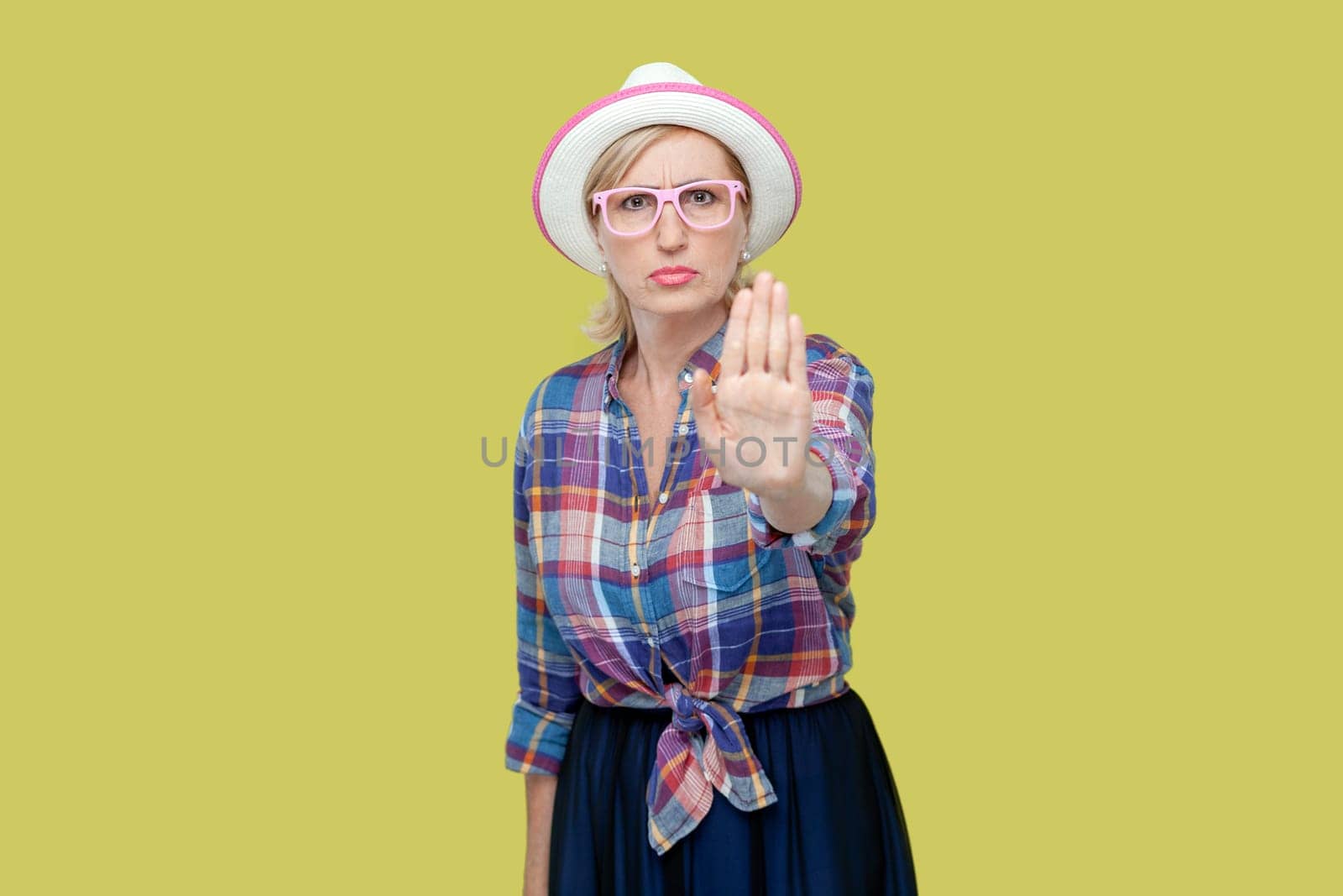 Portrait of serious senior woman wearing checkered shirt, hat and eyeglasses raises palm in stop or prohibition gesture, says no wait sec, hold on. Indoor studio shot isolated on yellow background