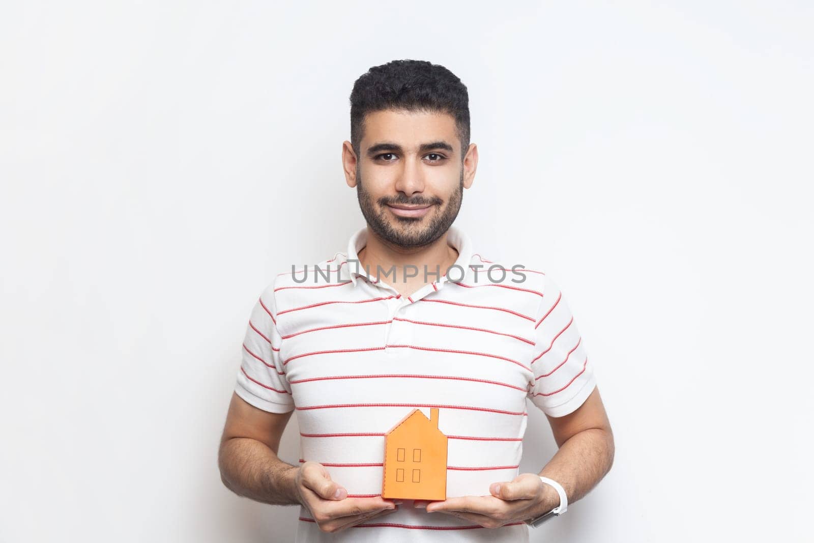 Portrait of attractive calm smiling bearded man wearing striped t-shirt standing holding paper house in hands, looking at camera, real estate agency. Indoor studio shot isolated on gray background.