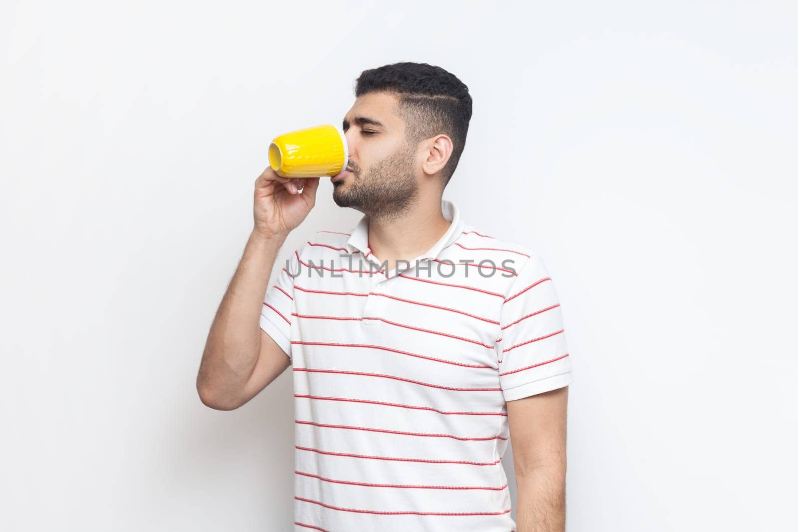 Portrait of handsome young adult bearded man wearing striped t-shirt standing holding yellow cup, drinking coffee, tea or water. Indoor studio shot isolated on gray background.