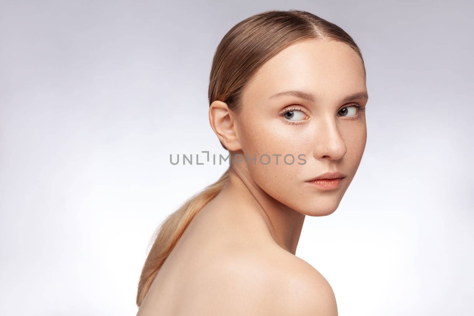 Side view portrait of serious pensive beautiful woman with natural makeupand ponytail, posing with bare shoulders, looking away. Indoor studio shot isolated over gray background.
