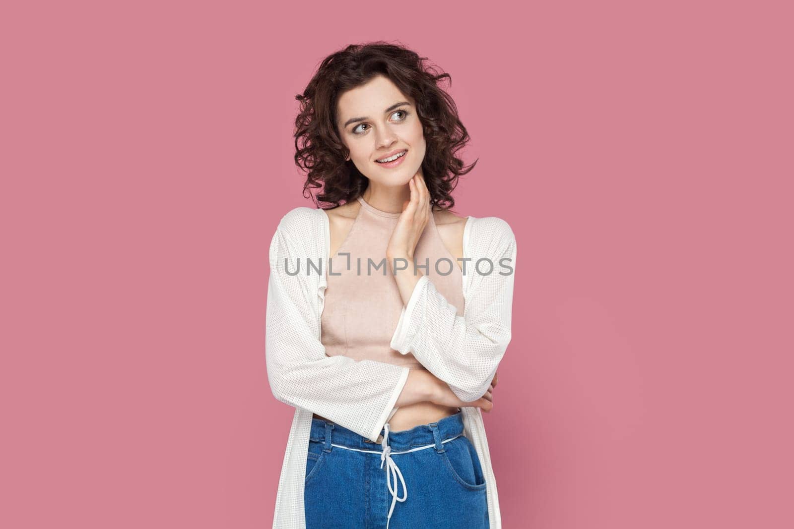 Portrait of friendly pleased smiling woman with curly hair wearing casual style outfit, looking away, dreaming about something pleasant. Indoor studio shot isolated on pink background.