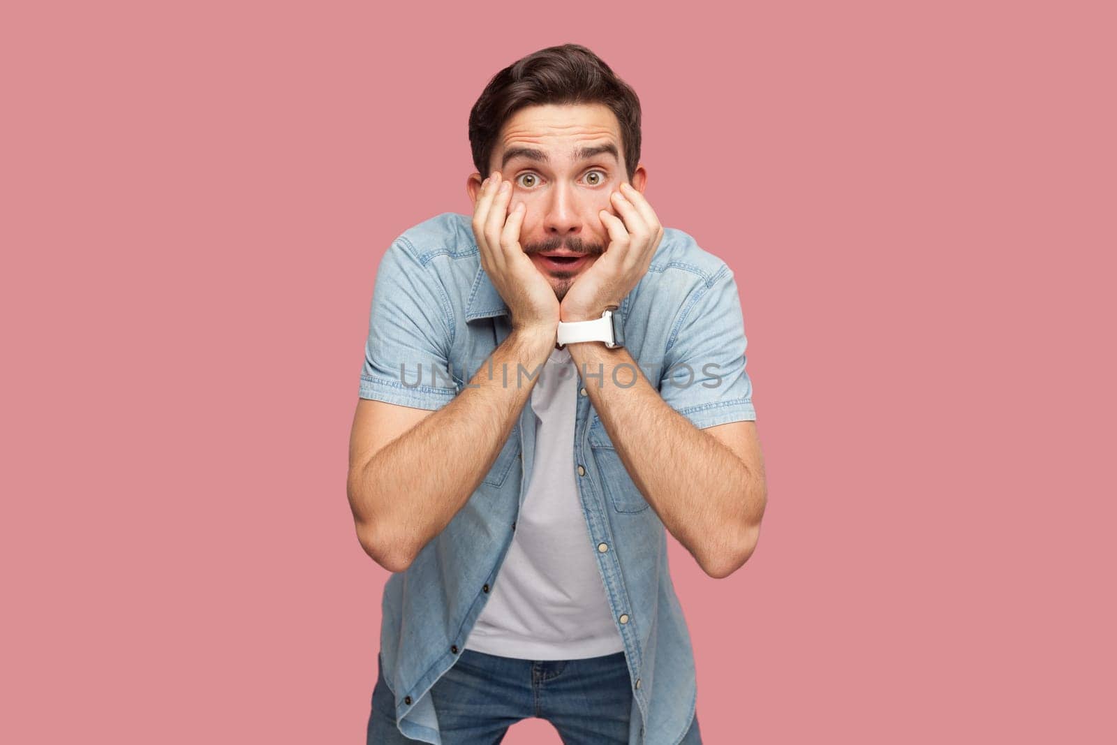 Portrait of shocked astonished bearded man in blue casual style shirt standing with hands on cheeks, looking at camera with big eyes. Indoor studio shot isolated on pink background.