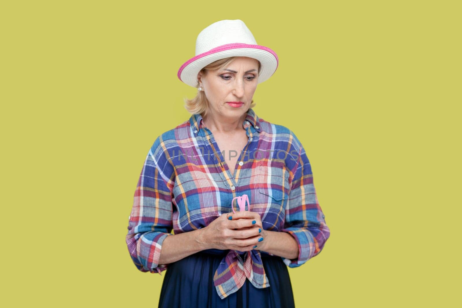 Unamused sad miserable mature woman wearing checkered shirt and hat, frowns face with dissatisfaction, sulks troubled as someone hurt her feelings. Indoor studio shot isolated on yellow background.