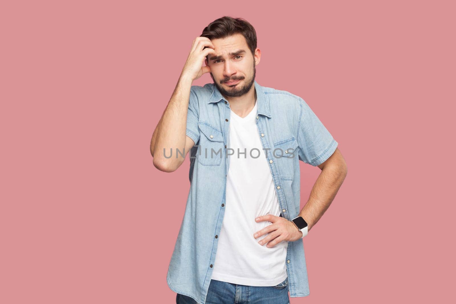 Portrait of puzzled bearded man in blue casual style shirt standing looks confusingly at camera, keeps hand on hip, rubbing his head. Indoor studio shot isolated on pink background.