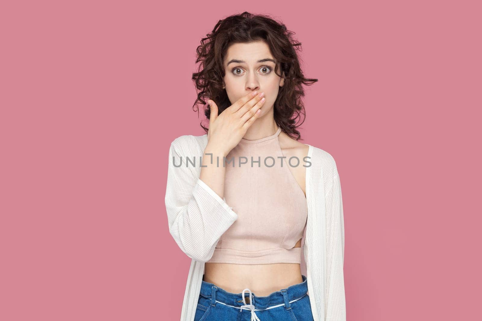 Portrait of shocked attractive woman with curly hair wearing casual style outfit covering mouth with palm, saying I won't tell anyone. Indoor studio shot isolated on pink background.