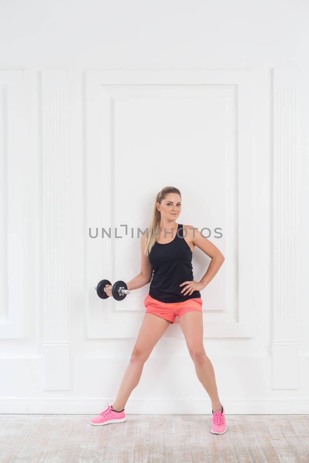 Full length portrait of young sporty confident beautiful woman in pink shorts and black top holding dumbbells doing exercise for arms at the gym on white wall background. Indoor studio shot.