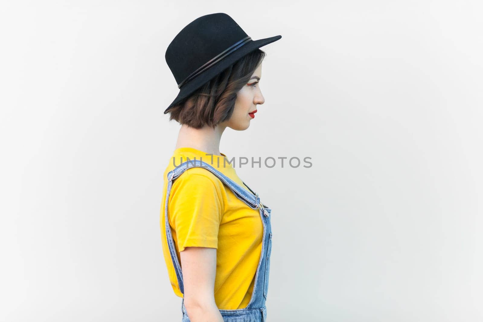 Side view portrait of serious hipster woman in denim overalls, yellow T-shirt and black hat, looking ahead, having strict expression, being in bad mood. Indoor studio shot isolated on gray background.