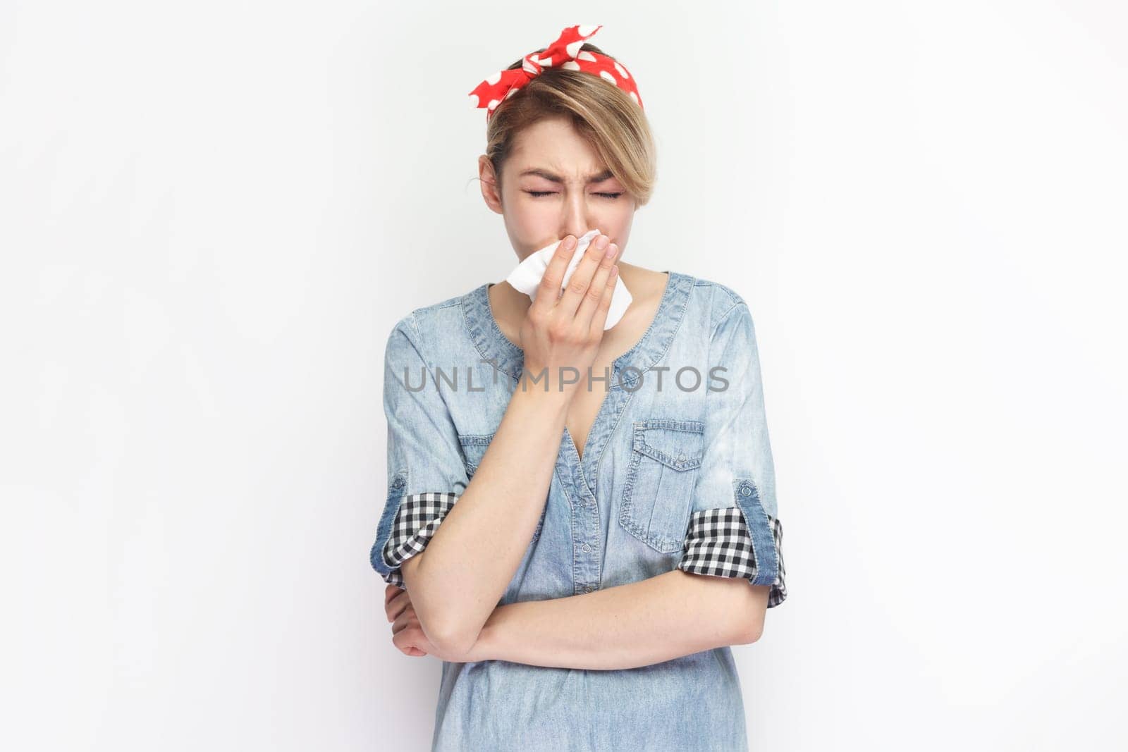 Portrait of sick unhealthy blonde woman wearing blue denim shirt and red headband standing suffering runny nose, having grippe symptoms. Indoor studio shot isolated on gray background.