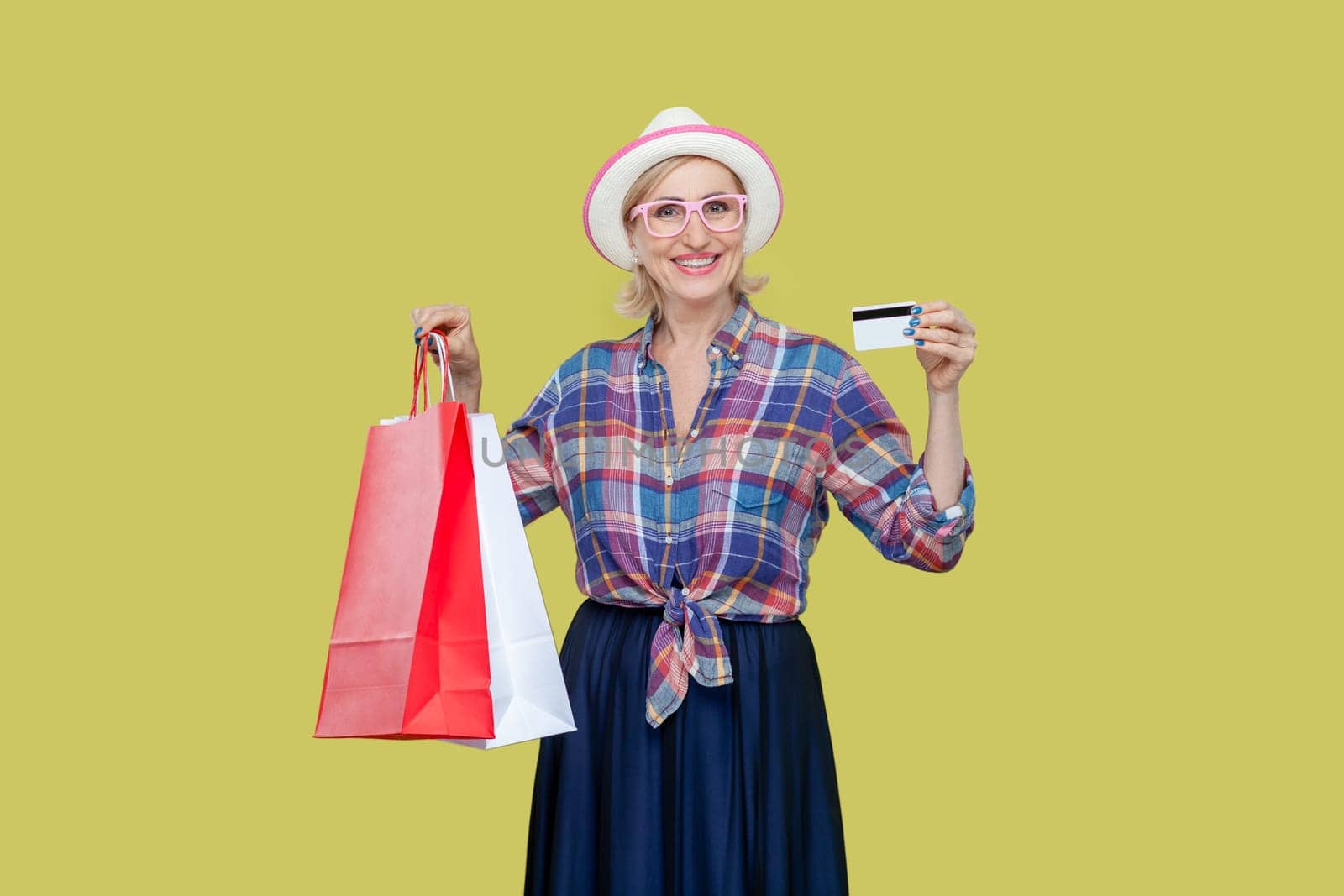 Portrait of smiling positive cheerful mature woman wearing checkered shirt, hat and eyeglasses holding shopping bag and showing credit card. Indoor studio shot isolated on yellow background.