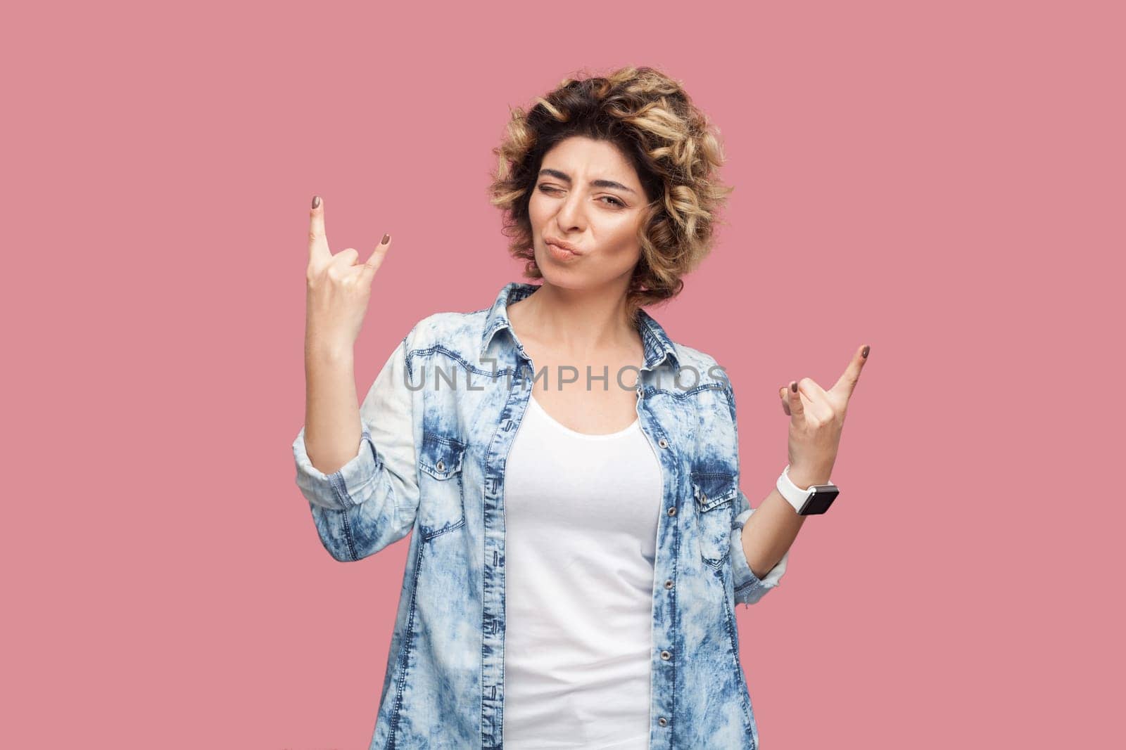 Portrait of cool festive woman with curly hairstyle wearing blue shirt standing and showing rock and roll gesture, enjoying concert. Indoor studio shot isolated on pink background.