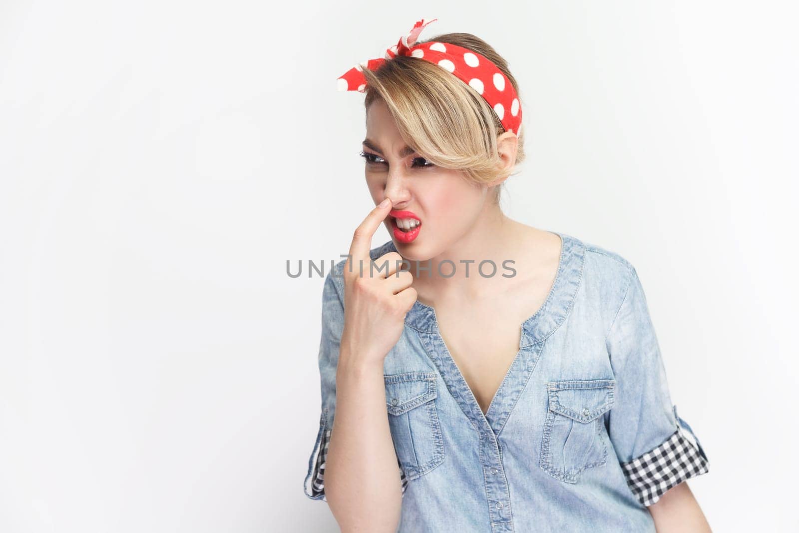 You are liar. Portrait of funny foolish blonde woman wearing blue denim shirt and red headband standing touching her nose, frowning face. Indoor studio shot isolated on gray background.