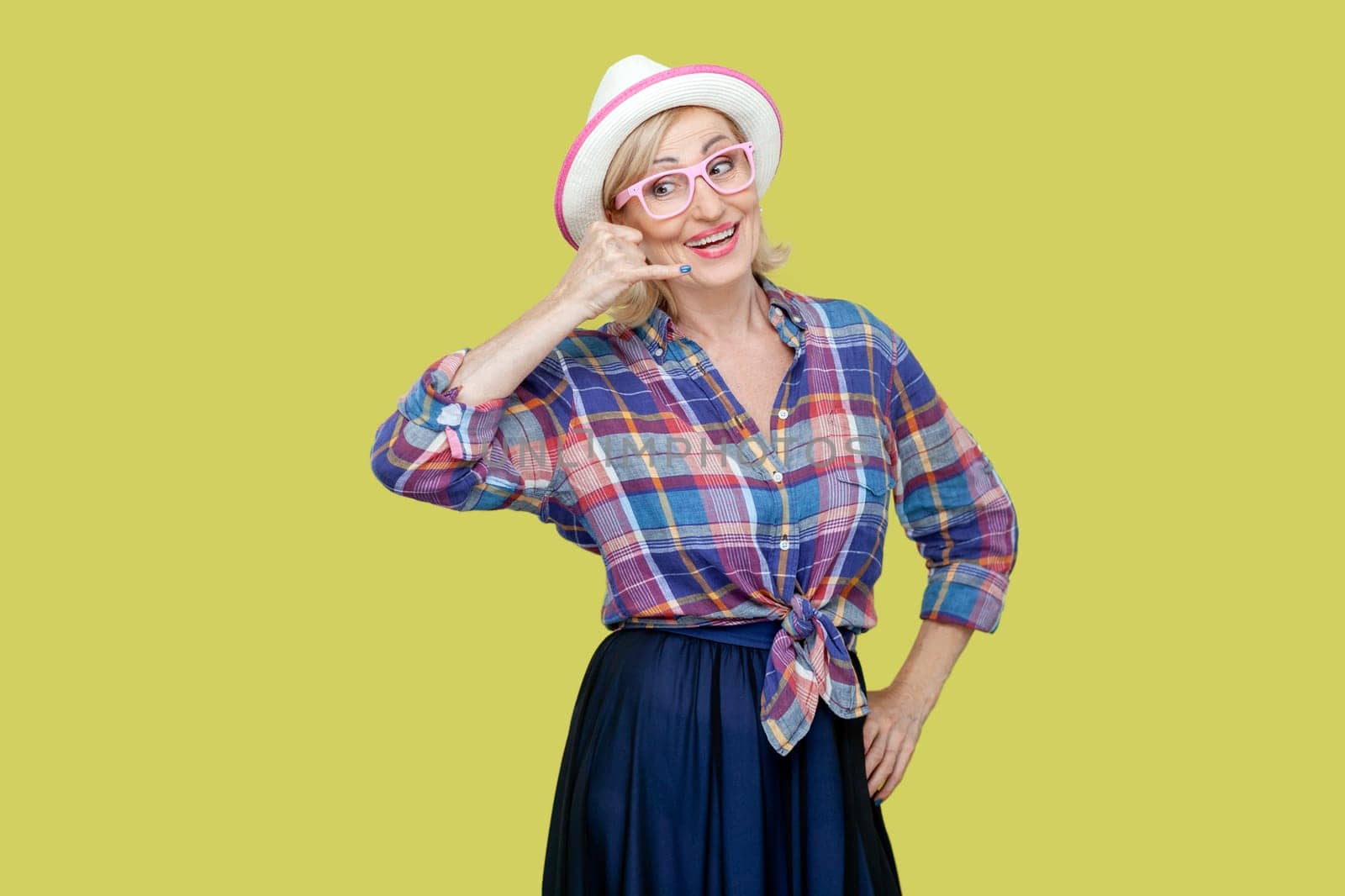 Portrait of positive cheerful senior woman wearing checkered shirt, hat and eyeglasses makes phone gesture, says call me back. Indoor studio shot isolated on yellow background.