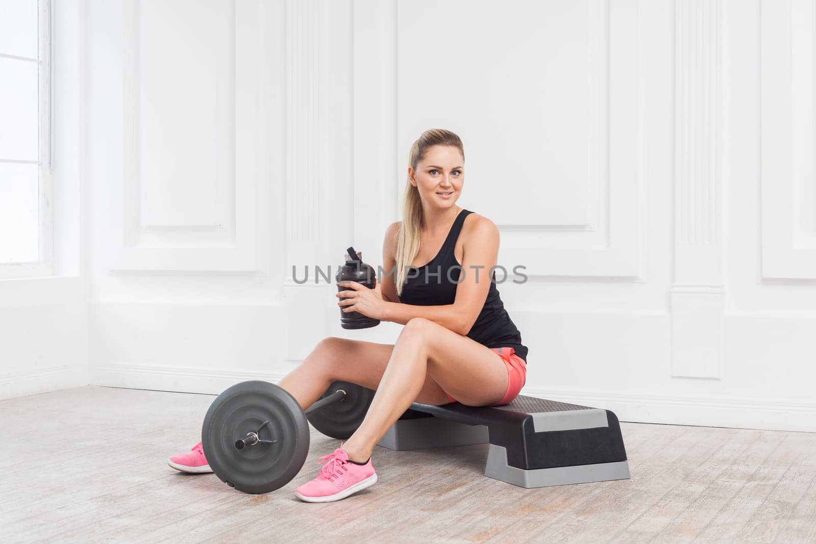 Portrait of happy smiling beautiful sporty woman wearing pink shorts and black top siting and have a rest after hard working on the gym, holding bottle with water. Indoor studio shot.