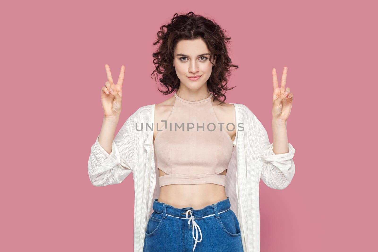 Portrait of attractive beautiful young adult woman with curly hair wearing casual style outfit standing looking at camera, showing v sign. Indoor studio shot isolated on pink background.