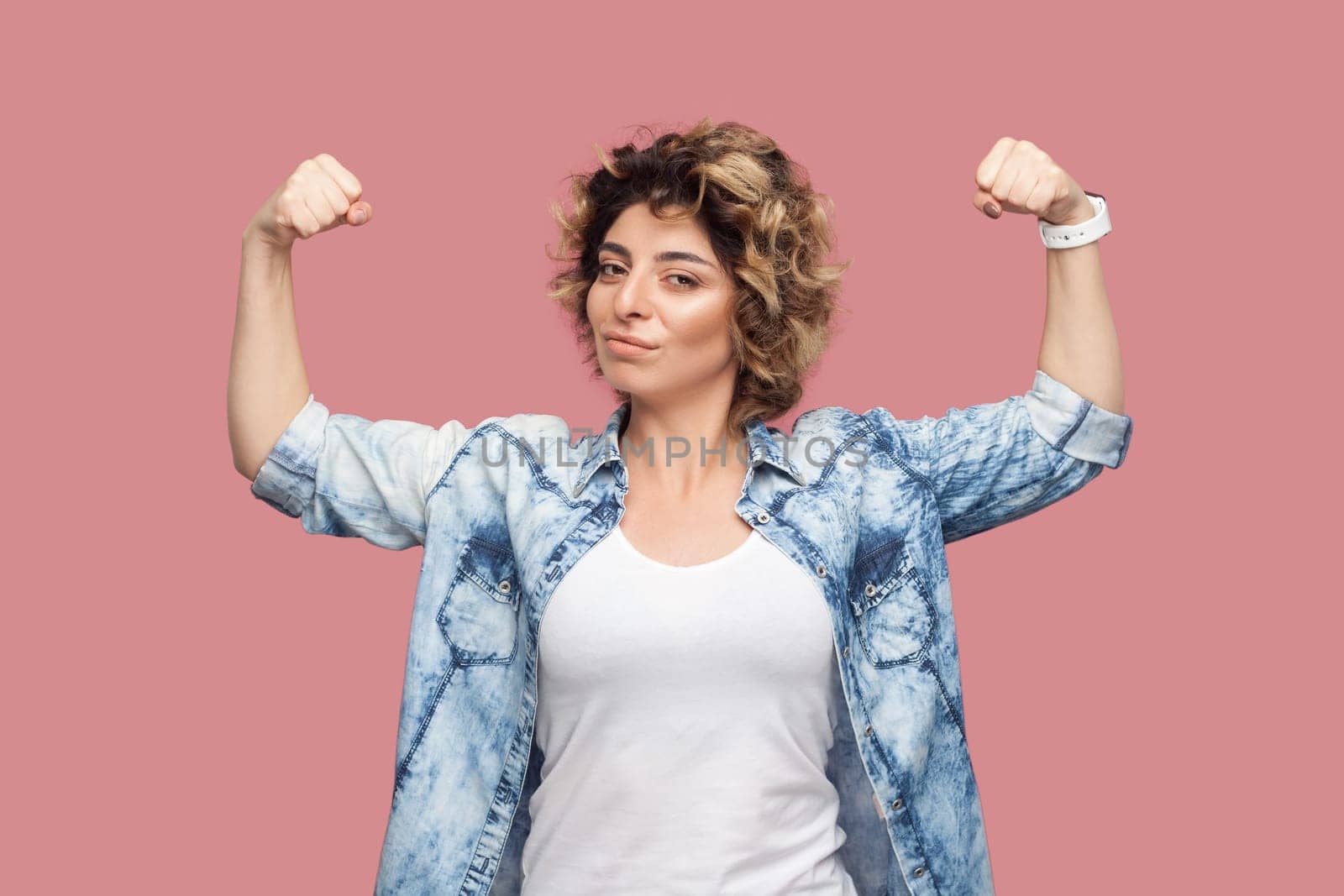Portrait of self confident strong woman with curly hairstyle wearing blue shirt standing with raised arms, showing her power, looking at camera. Indoor studio shot isolated on pink background.