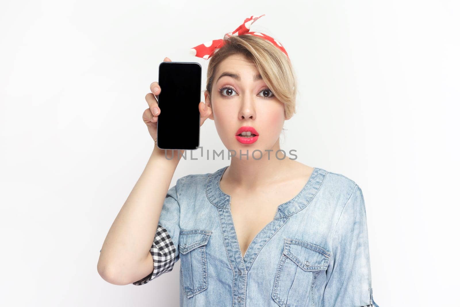 Portrait of shocked blonde young woman wearing blue denim shirt and red headband standing holding smart phone with black blank screen. Indoor studio shot isolated on gray background.