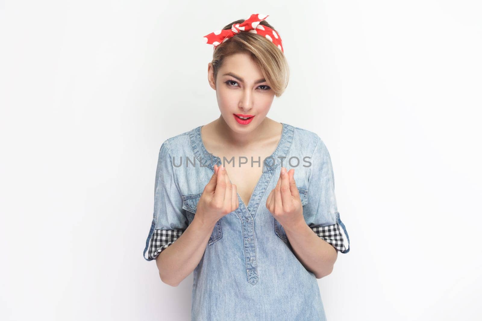 Portrait of attractive beautiful blonde woman wearing blue denim shirt and red headband standing showing money gesture with fingers. Indoor studio shot isolated on gray background.