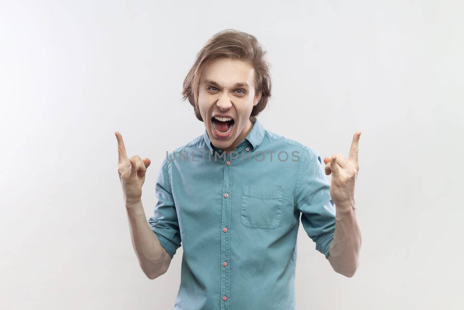 Portrait of crazy excited young man having fun at rock festival, showing rock and roll gesture, screaming, yelling, wearing blue shirt. Indoor studio shot isolated on gray background.