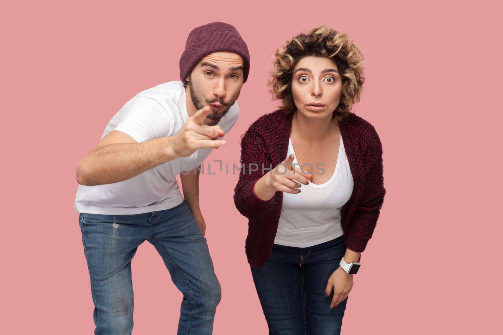 Portrait of astonished couple of friends standing together and pointing at camera, choosing you, looking with big eyes, being amazed. Indoor studio shot isolated on pink background.