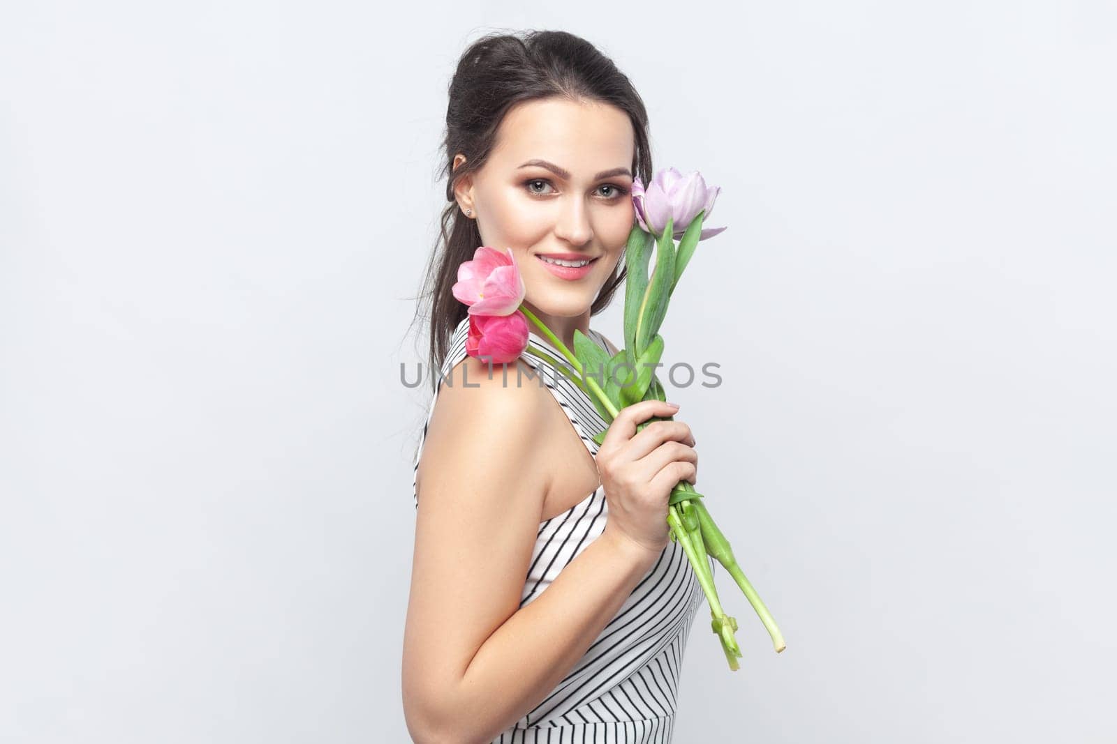 Portrait of smiling beautiful brunette woman holding bouquet of tulips, looking at camera, smiling with happiness, wearing striped dress. Indoor studio shot isolated on gray background.