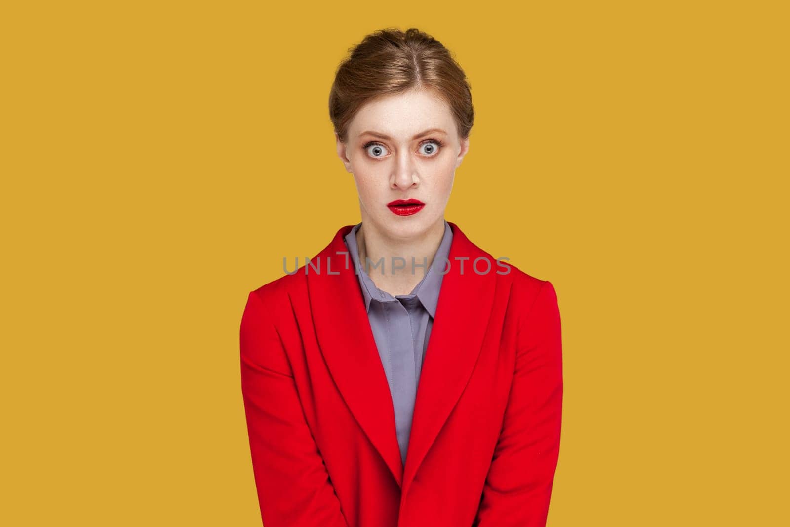 Portrait of shocked scared frighten woman with red lips standing looking at camera with big eyes, sees something scary, wearing red jacket. Indoor studio shot isolated on yellow background.