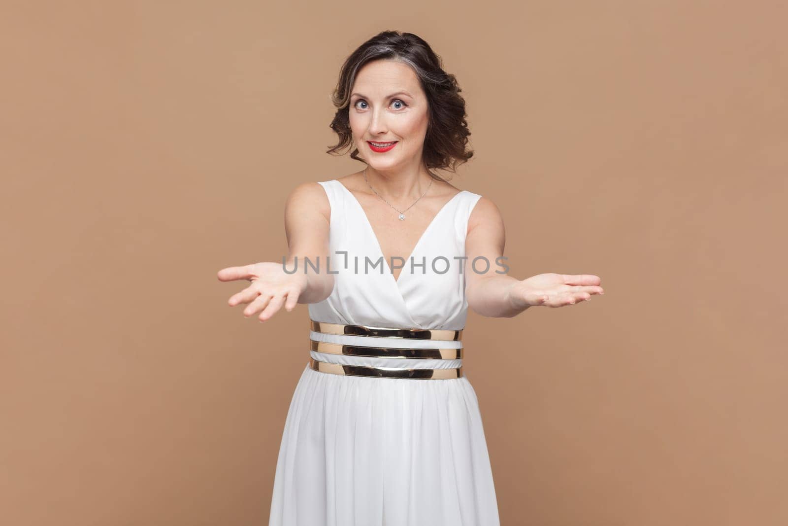 Please, take for free. Kind generous woman outstretching hands as if giving for free, offering to embrace, inviting with hospitable expression. Indoor studio shot isolated on light brown background.