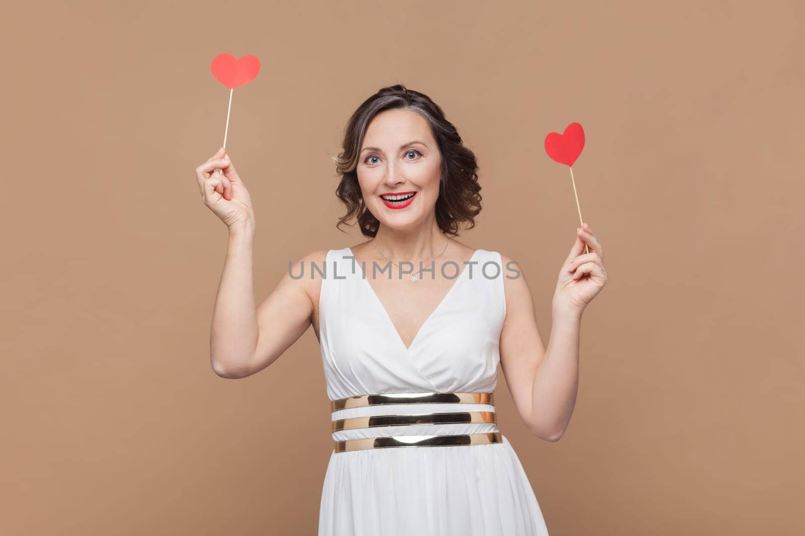 Portrait of excited positive middle aged woman with wavy hair holding little red hearts, smiling happily, celebrating, wearing white dress. Indoor studio shot isolated on light brown background.