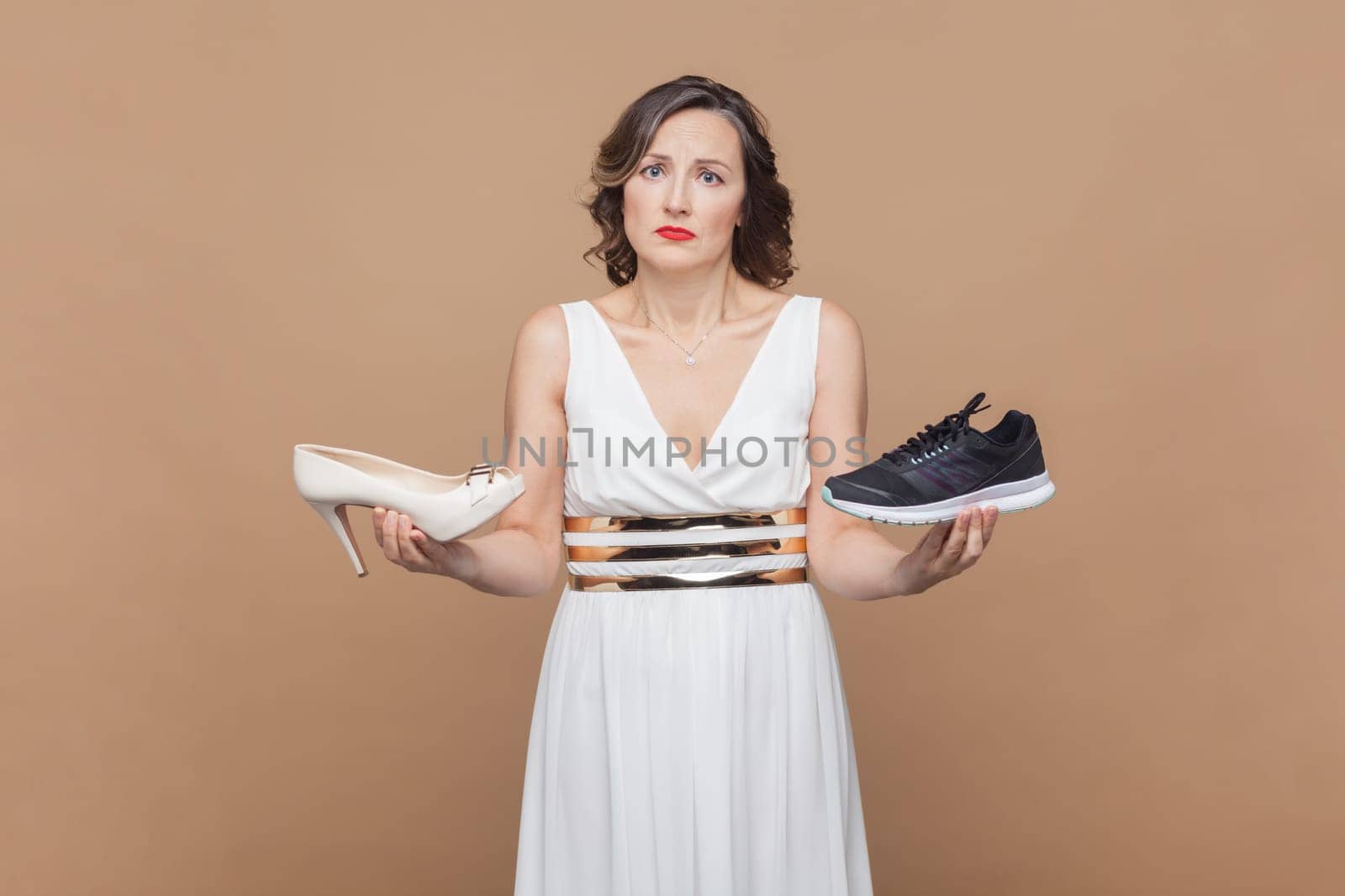 Portrait of sad doubtful middle aged woman with wavy hair holding sneakers and high heels, looking at camera with puzzled face, make decision. Indoor studio shot isolated on light brown background.
