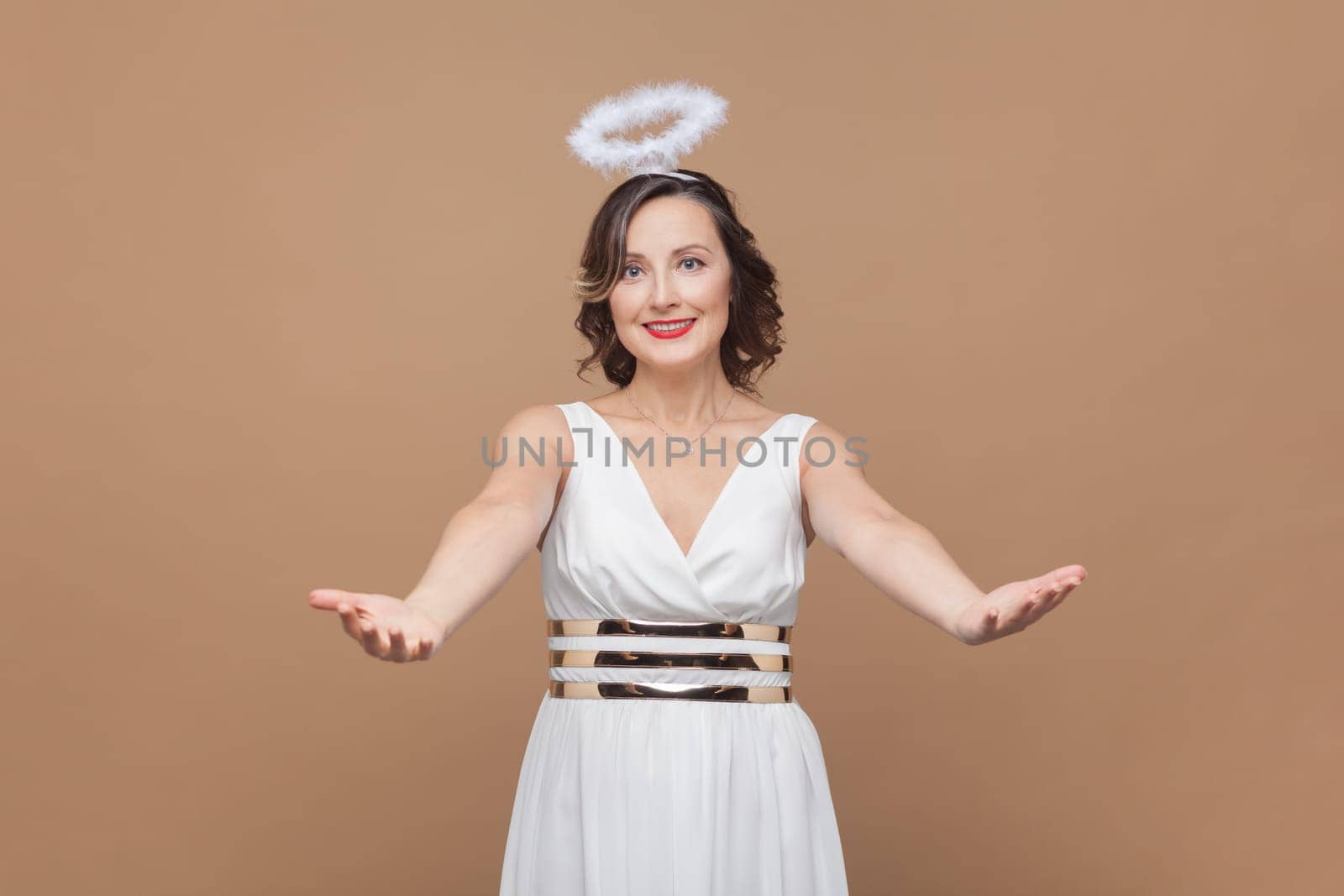 Free hugs, come into my arms. Woman with wavy hair and nimb over head, stretching hands to camera, smiling broadly, going to embrace, share love. Indoor studio shot isolated on light brown background.