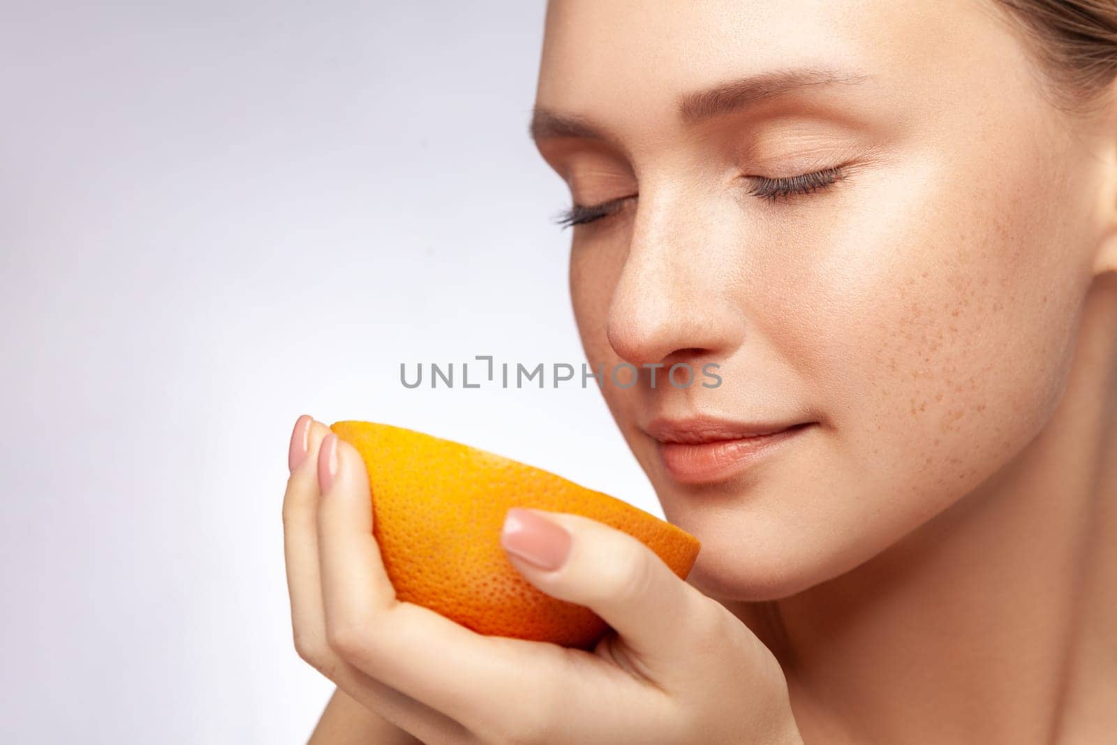 Closeup portrait of dreamy beautiful woman with closed eyes holding smelling half of grapefruit, model with natural makeup. Indoor studio shot isolated over gray background.
