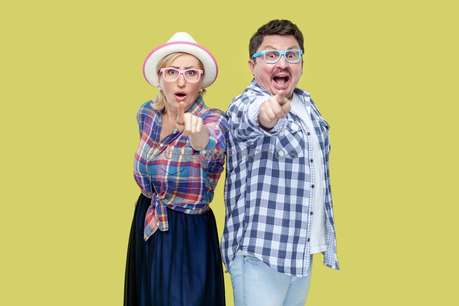Shocked couple of friends, adult man and woman in casual checkered shirt standing together and pointing at camera, expressing astonishment. Indoor studio shot isolated, on yellow background.