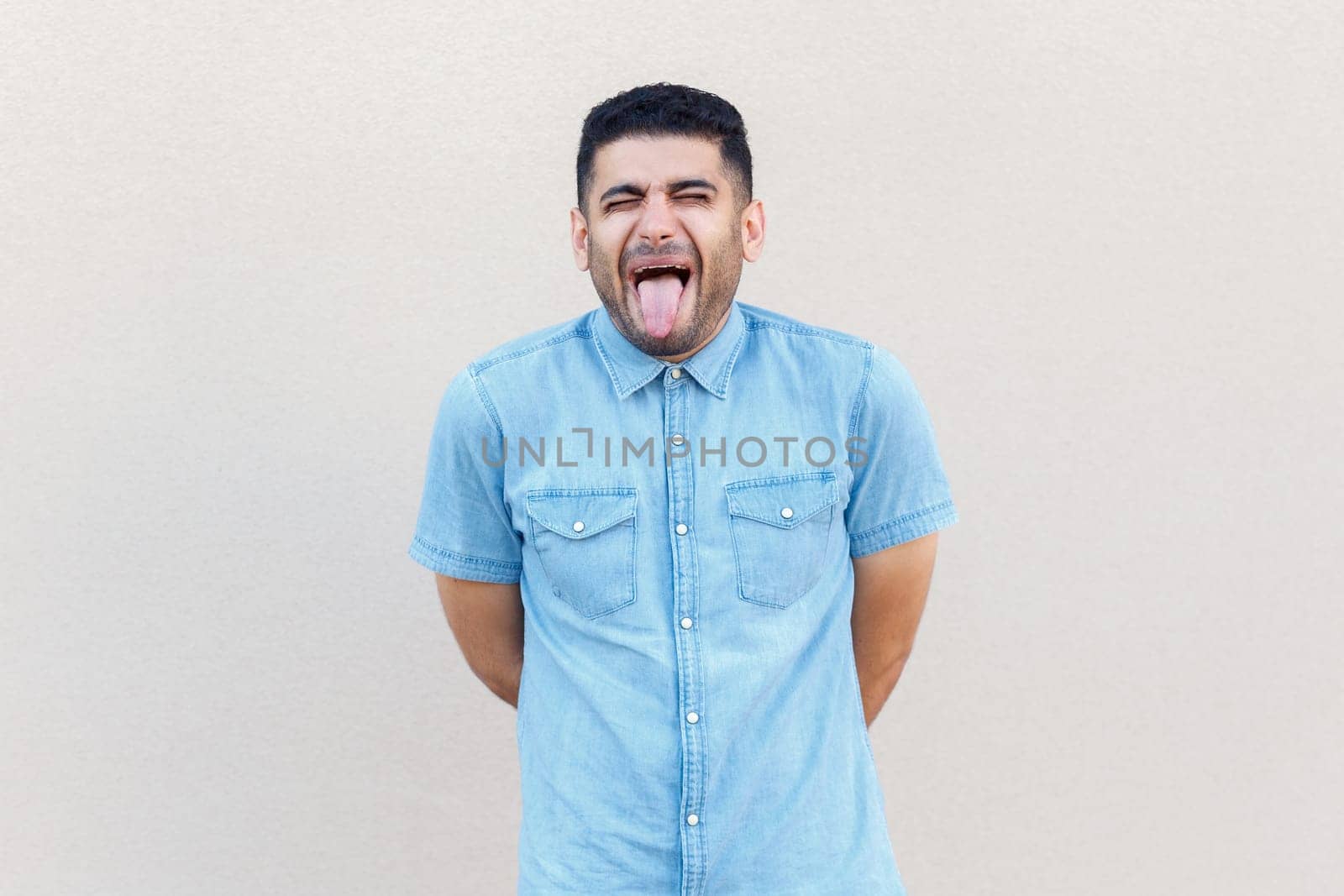 Portrait of funny crazy handsome man wearing denim shirt standing with closed eyes and showing tongue out, demonstrates childish behavior. Indoor studio shot isolated on gray background.
