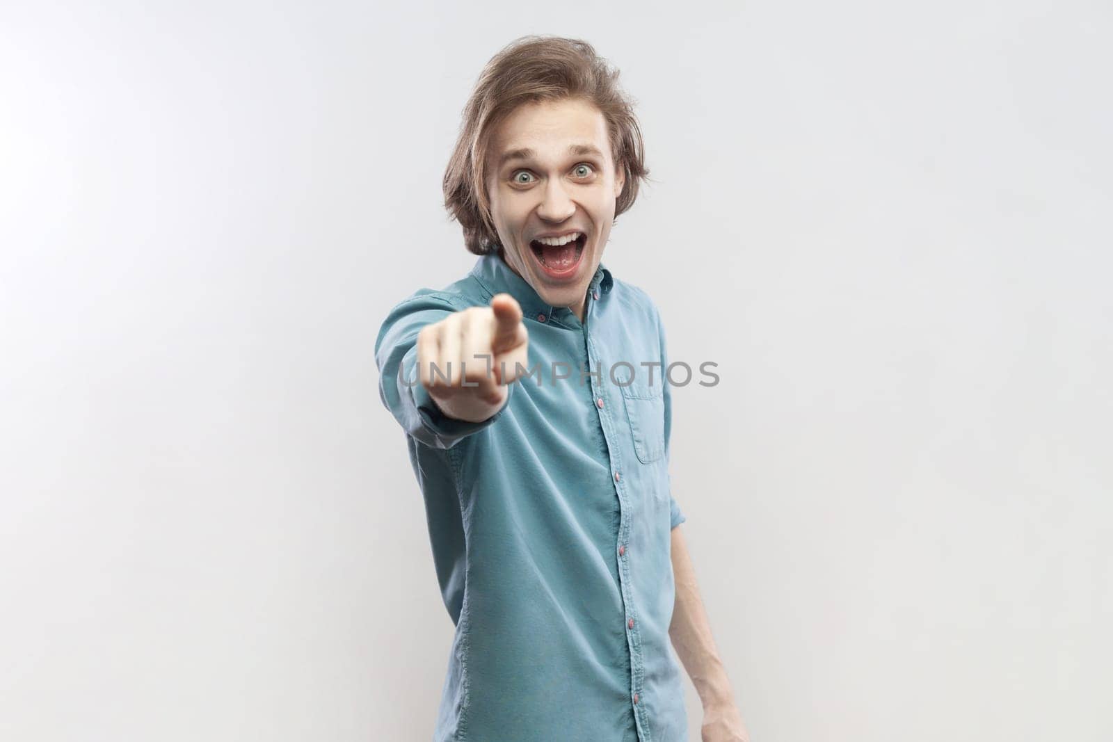 Portrait of surprised astonished shocked young man standing pointing at camera, selecting you, expressing positive emotions, wearing blue shirt. Indoor studio shot isolated on gray background.